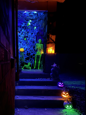 A blacklight Haloween scene. Th back drop is spiderwebs, there is a glowing Jackolantern on a stand, glowing bats hung from the ceiling, a spider on the webs, a green crawling skeleton and a green glowing skeleton waving hello. On the edge of the steps are 3 jackolanterns and a gargoyle. A few candles are around.