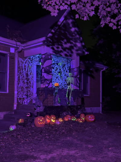 A blacklight Haloween scene. Th back drop is spiderwebs, there is a glowing Jackolantern on a stand, glowing bats hung from the ceiling, a spider on the webs, a green crawling skeleton and a green glowing skeleton waving hello. On the edge of the steps are 3 jackolanterns and a gargoyle. A few candles are around. Beside the porch is a row of Jackolanterns lit in different colors.