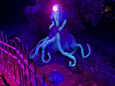 A lampreyfaced octomermaid in a 1980's style prom dress. Magenta light comes out of her face as she stands next to the graveyard fence. 